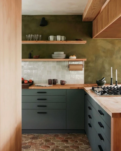 Modern kitchen interior with green cabinets, wooden countertops, open shelves, and brick flooring.