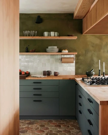Modern kitchen interior with green cabinets, wooden countertops, open shelves, and brick flooring.