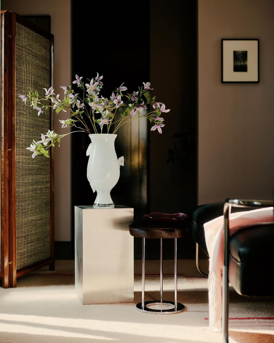 Interior design featuring a white vase with lilac flowers atop a silver pedestal table, with a screen and chair in the background.
