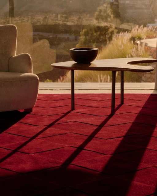 Living room interior showcasing a modern coffee table with a bowl, soft armchair, and a red patterned rug near a bright window.