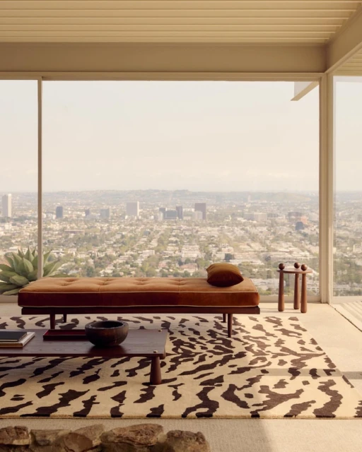 Daybed with pillow and coffee table on an animal print rug inside a modern house with a city view through large windows.