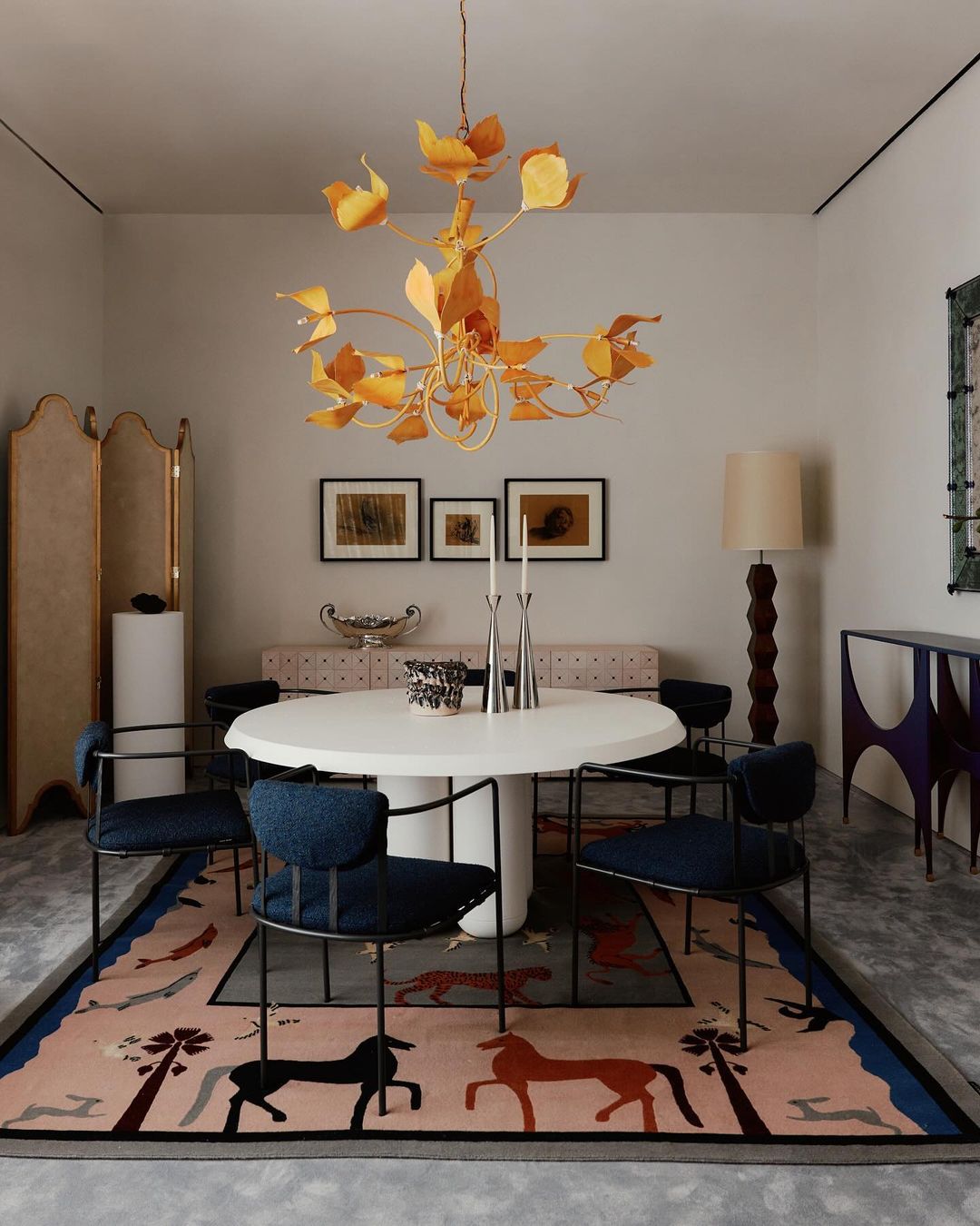 Indoor dining room scene with a white round table, blue chairs, a golden chandelier, and a patterned rug featuring horses and trees.
