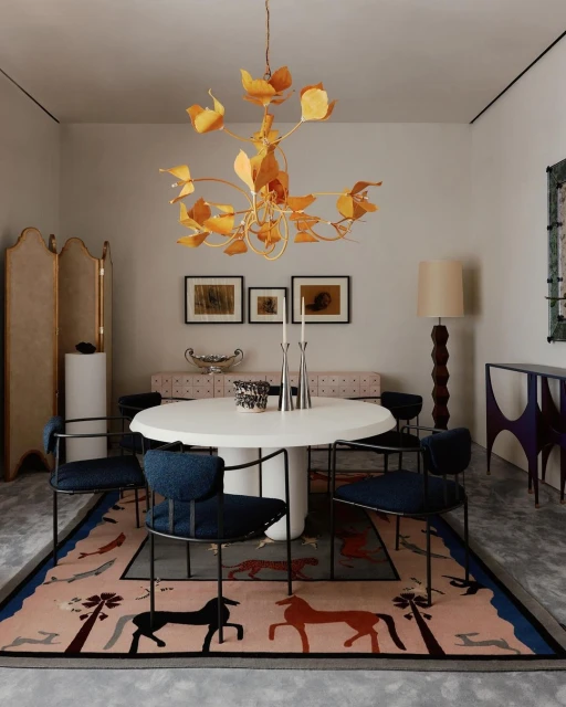 Indoor dining room scene with a white round table, blue chairs, a golden chandelier, and a patterned rug featuring horses and trees.