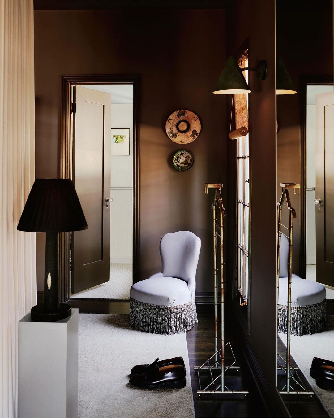 Interior view of a sitting room with a chair, shoes, a mirror, a lamp on a pedestal, and decorative plates against dark brown walls.