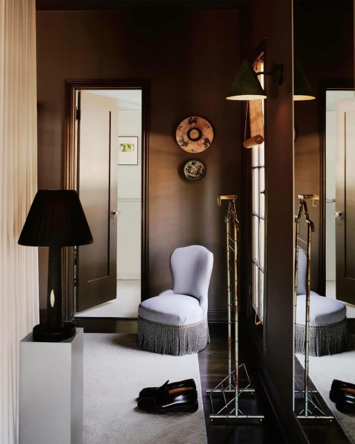 Interior view of a sitting room with a chair, shoes, a mirror, a lamp on a pedestal, and decorative plates against dark brown walls.