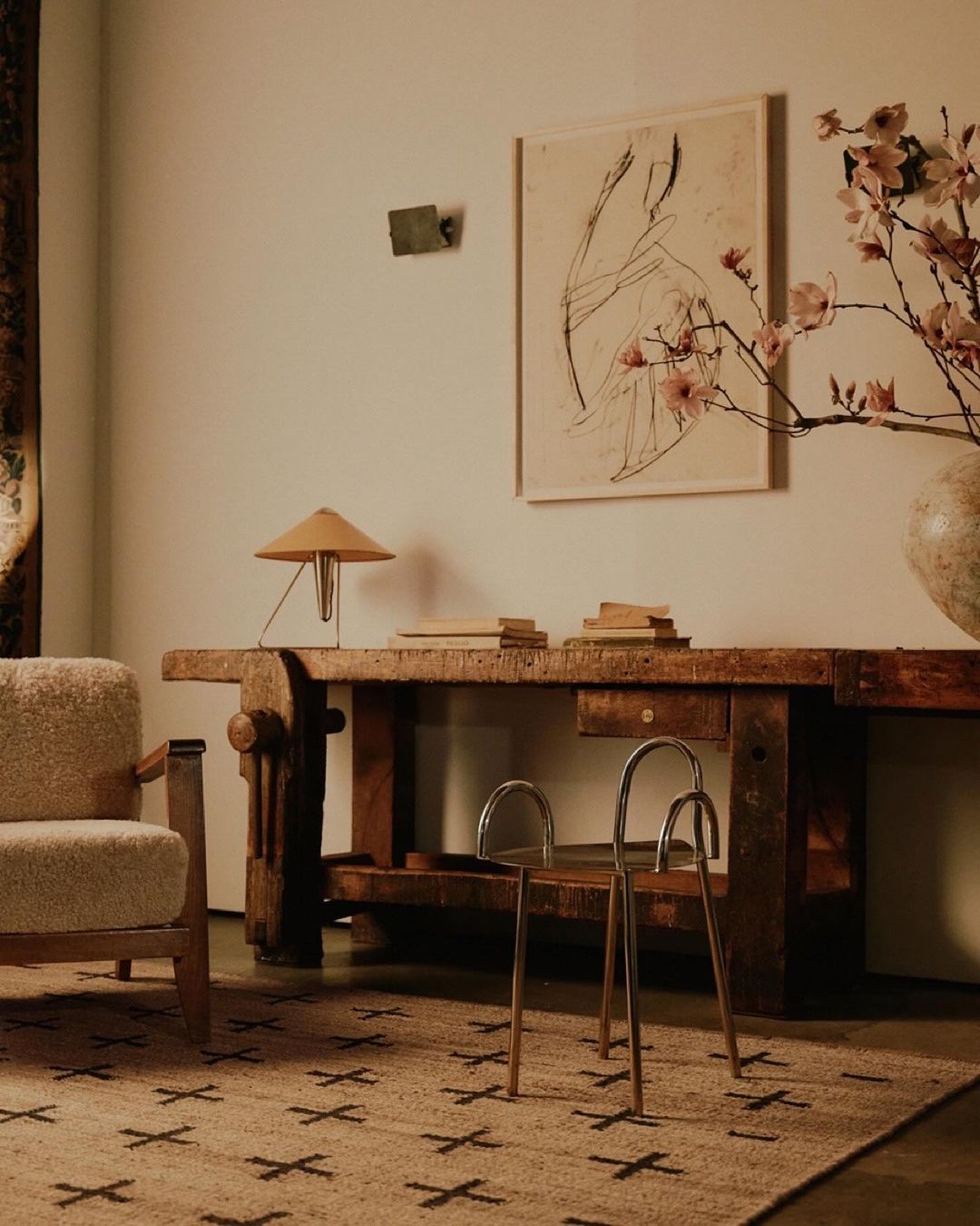 Interior shot of a room featuring a light wood table, chair, artwork, lamp, and patterned rug, bathed in warm, neutral tones and diffused light.