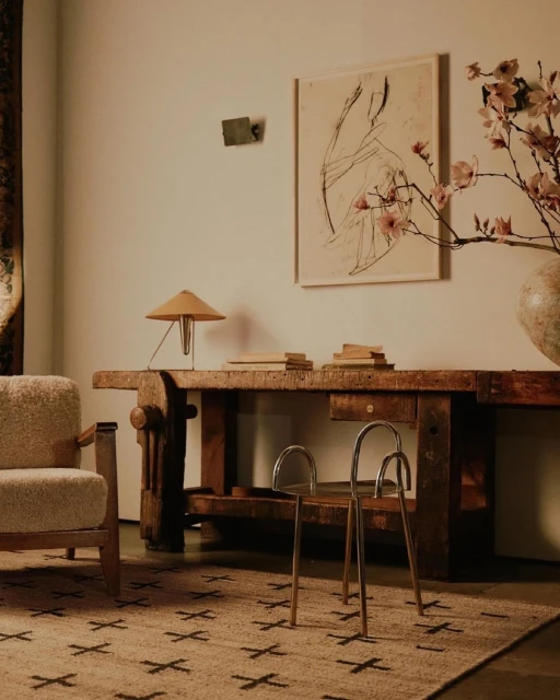 Interior shot of a room featuring a light wood table, chair, artwork, lamp, and patterned rug, bathed in warm, neutral tones and diffused light.