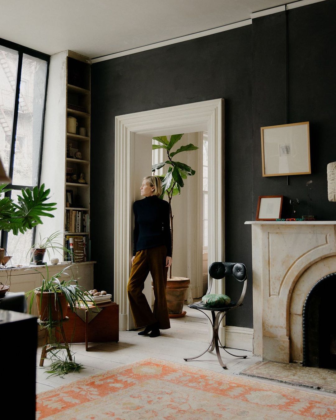 Woman in black turtleneck and brown pants stands in an elegant living room with neutral colors, plants, and a fireplace, gazing out a window.