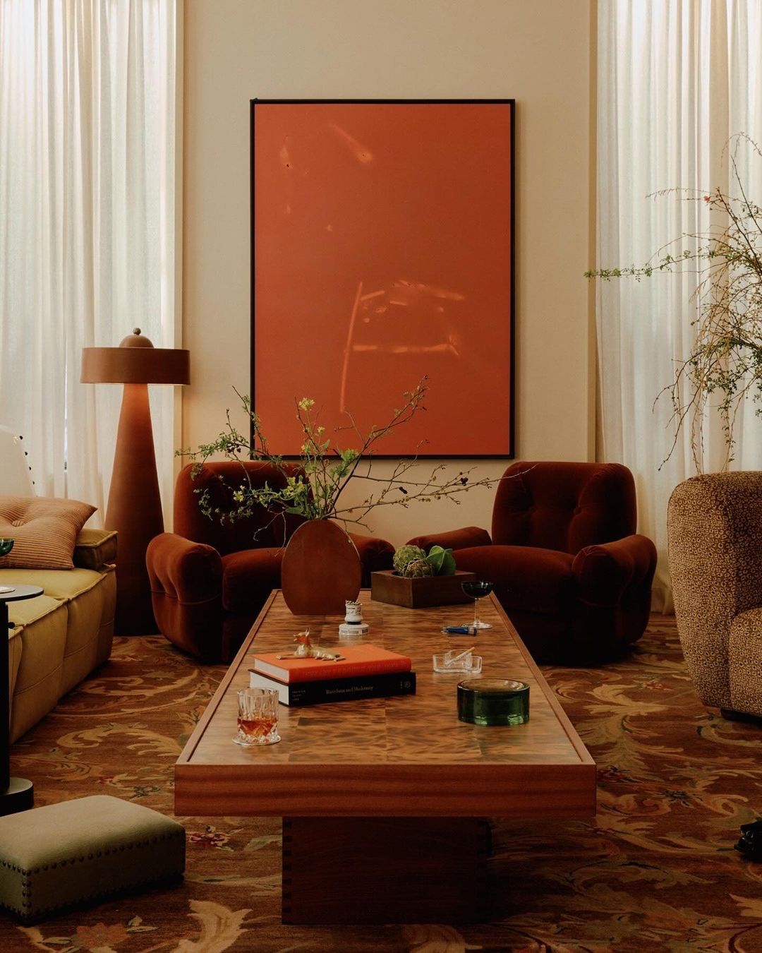 Living room interior with two rust-colored armchairs, a light-brown sofa, and a wood coffee table, illuminated by a lamp and natural light.