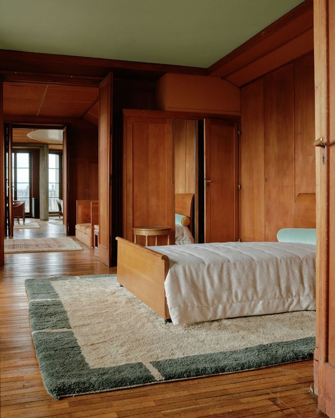Interior view of a bedroom with wood paneled walls, bed with white bedding, and a green and white rug on a wood floor.
