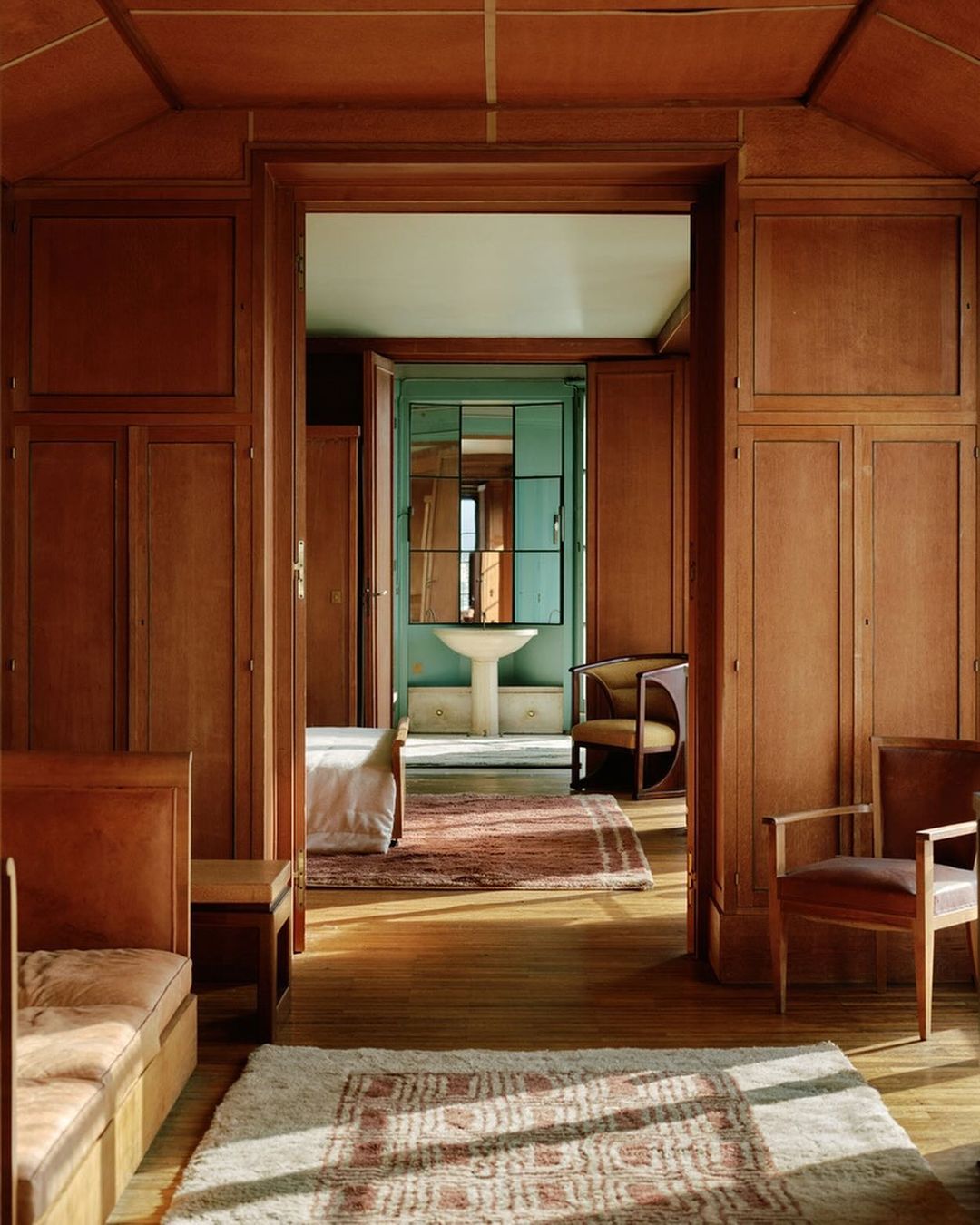 Classic interior view through a doorway showing a bedroom and a bathroom. Wood paneled walls and patterned rugs add to the traditional style.