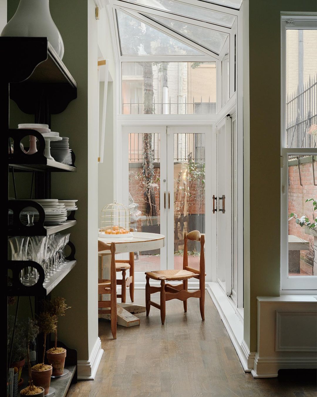 Well-lit dining area with a round table, wooden chairs, and a glass-roofed extension providing natural light, plus a decorative shelf.