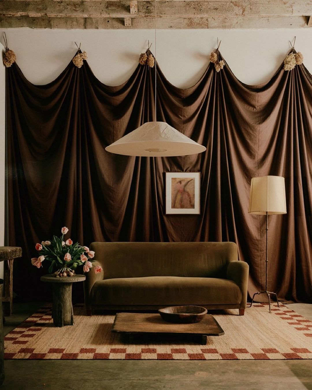 Living room interior featuring a green velvet sofa, round side table with flowers, brown wooden coffee table, and decorative brown drapes.