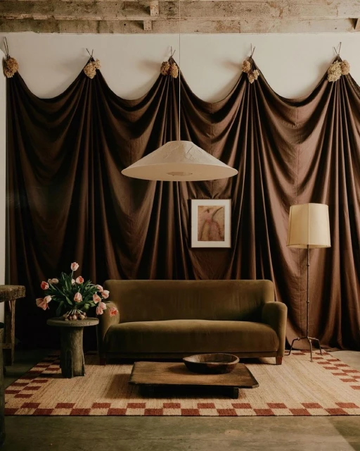 Living room interior featuring a green velvet sofa, round side table with flowers, brown wooden coffee table, and decorative brown drapes.