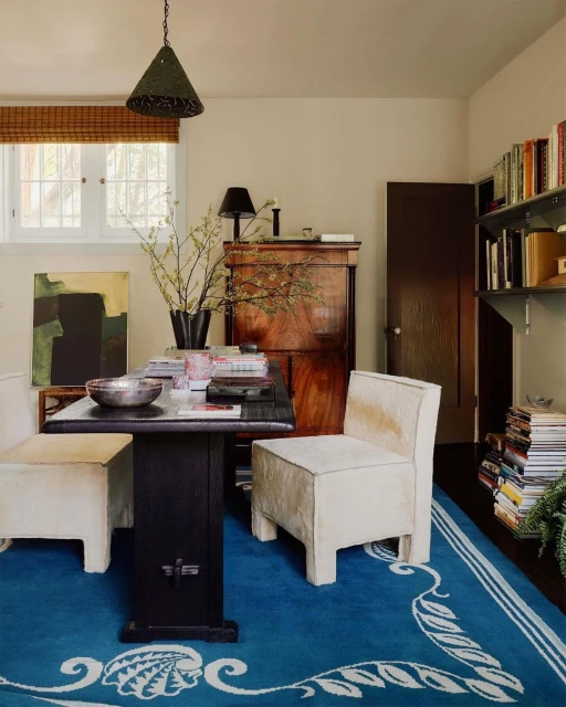 Cozy interior with a blue patterned rug, dark wood table, and upholstered chairs, illuminated by natural light and a hanging lamp.