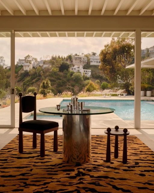 Indoor view of dining set with round table, chair, and tiger-print rug, looking out to a swimming pool and hillside houses.