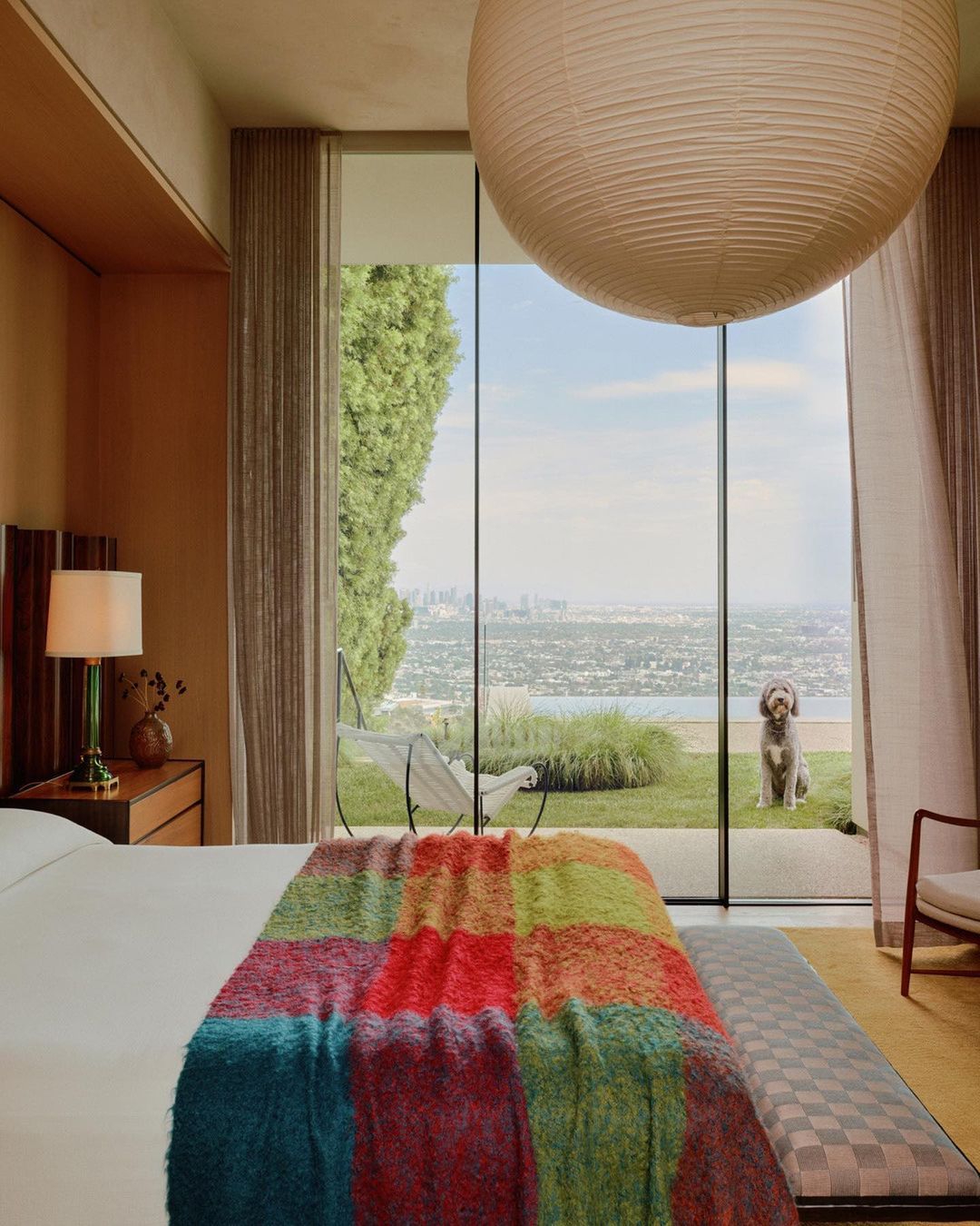 Bedroom interior with a colorful blanket, a green lawn, a city view and a dog, through large windows in daylight. 