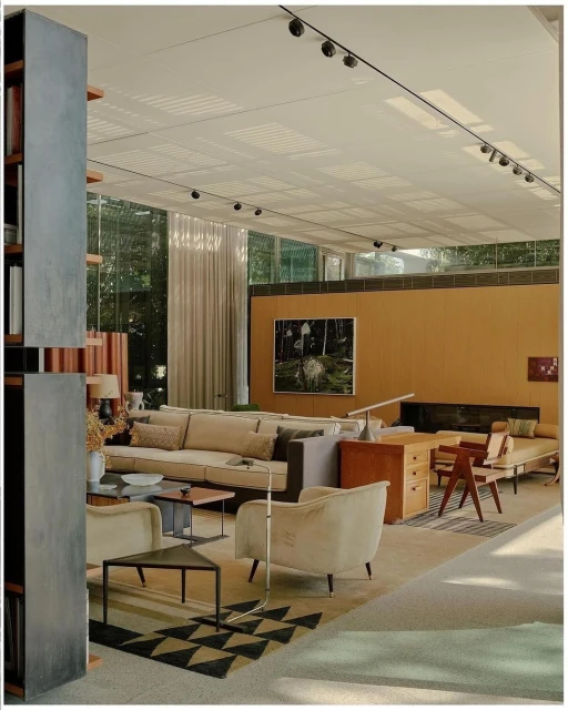 Sofa and armchairs arranged in a living room with triangle patterned rug, open to the outdoors via large glass windows, light neutral tones.