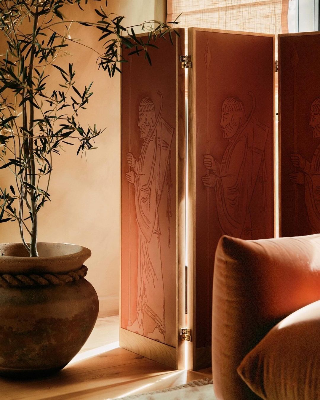 Orangey-tan interior with a potted plant, a decorative folding screen featuring line art of robed figures, and a plush cushion.