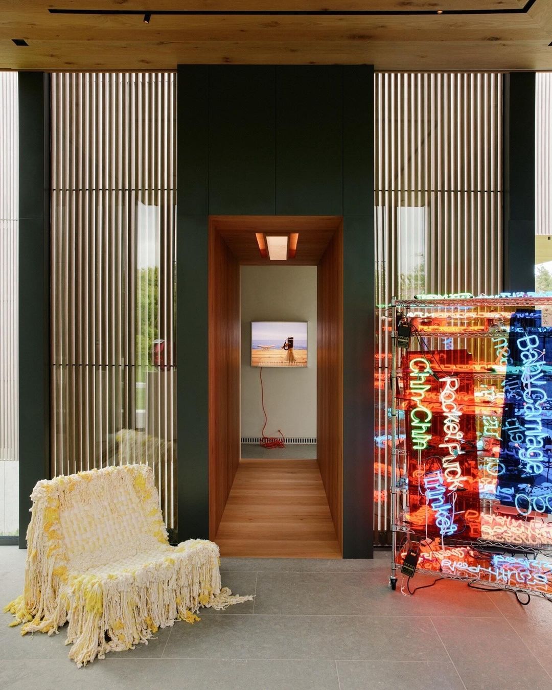Interior view of modern home with bright neon art, a shaggy chair, and a view into another room with a wall-mounted television.