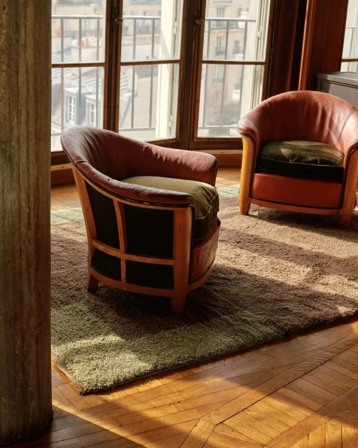 Two vintage armchairs sit on a green rug inside a room with natural light streaming through a large window showcasing a city view.