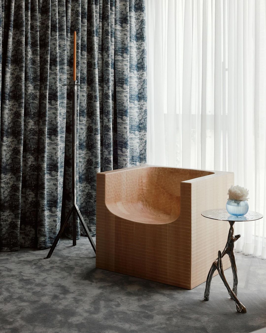 Interior design featuring a modern, light wood chair, round table with flowers, and patterned curtains. Natural light fills the room.
