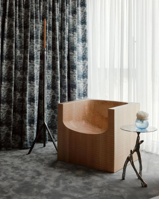 Interior design featuring a modern, light wood chair, round table with flowers, and patterned curtains. Natural light fills the room.