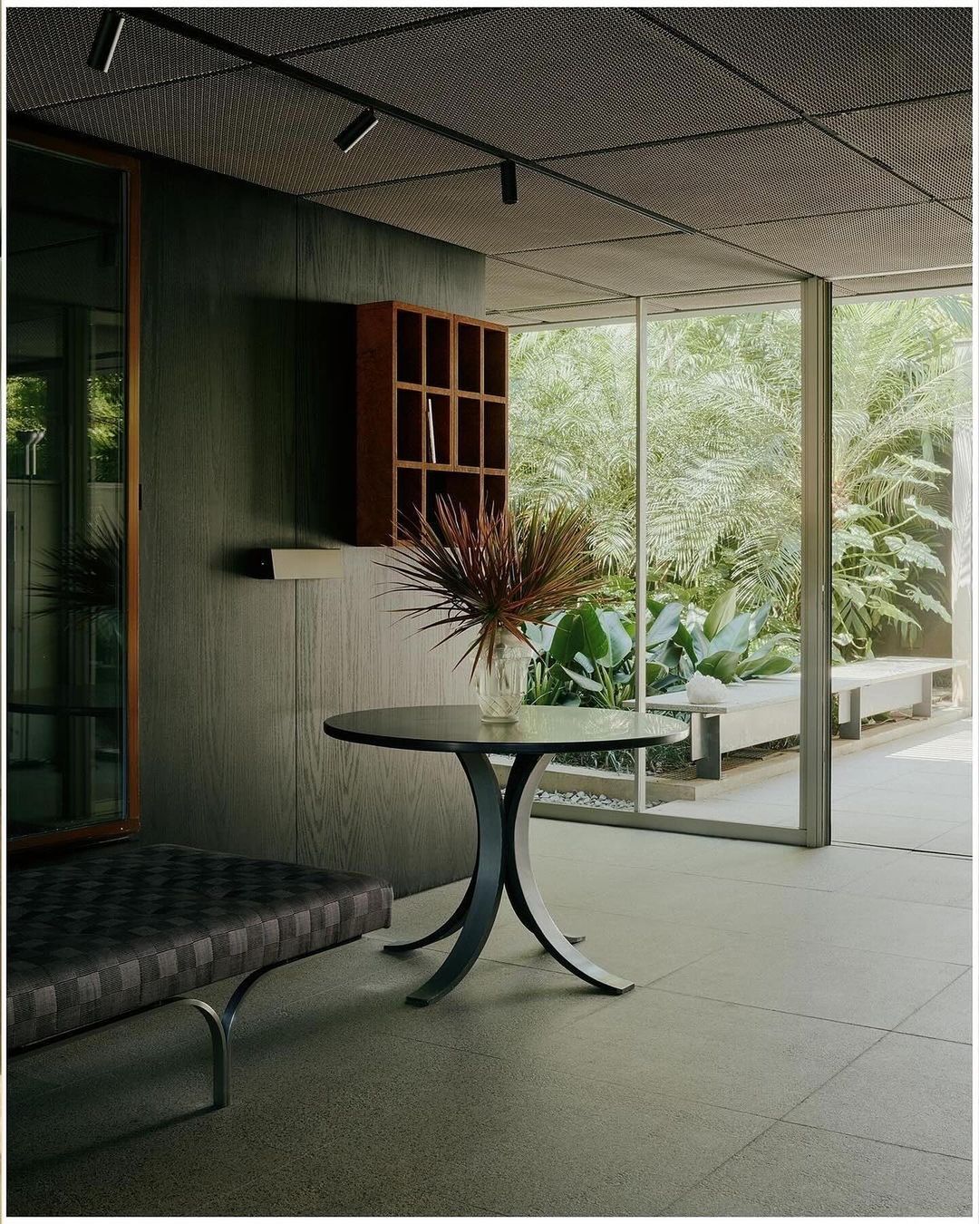 Indoor setting featuring a round table with a vase, a grey bench, a wooden shelf with compartments, and a view to an outdoor garden.