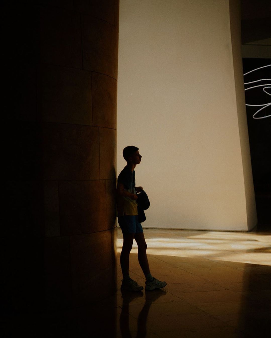 Silhouette of a young man leaning against a pillar, bag in hand, in a sunlit indoor space with light reflecting on the shiny floor.
