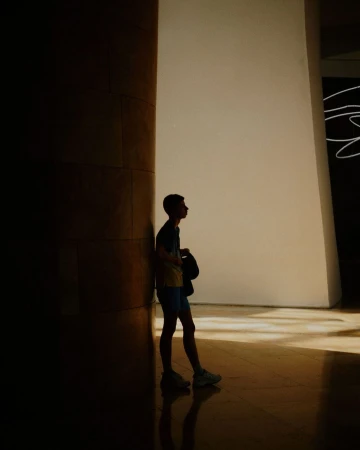 Silhouette of a young man leaning against a pillar, bag in hand, in a sunlit indoor space with light reflecting on the shiny floor.
