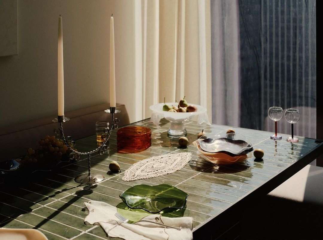 Indoor tablescape featuring a candle holder with lit candles, glass dishes, fruit, and draped white cloth, illuminated by natural lighting.