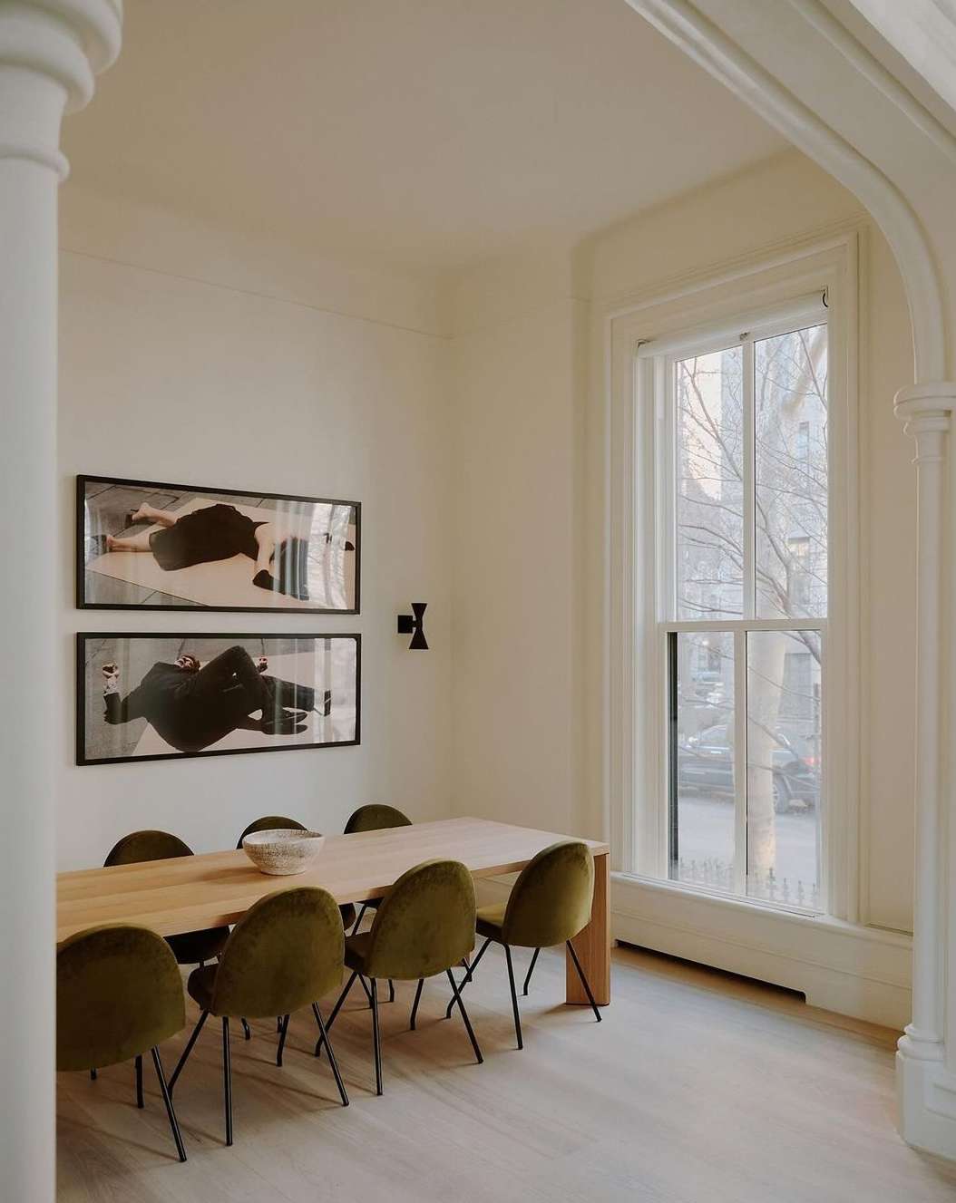 Dining room interior featuring a wooden table with olive green chairs, artwork on the wall, and natural light from a large window.