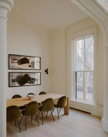 Dining room interior featuring a wooden table with olive green chairs, artwork on the wall, and natural light from a large window.