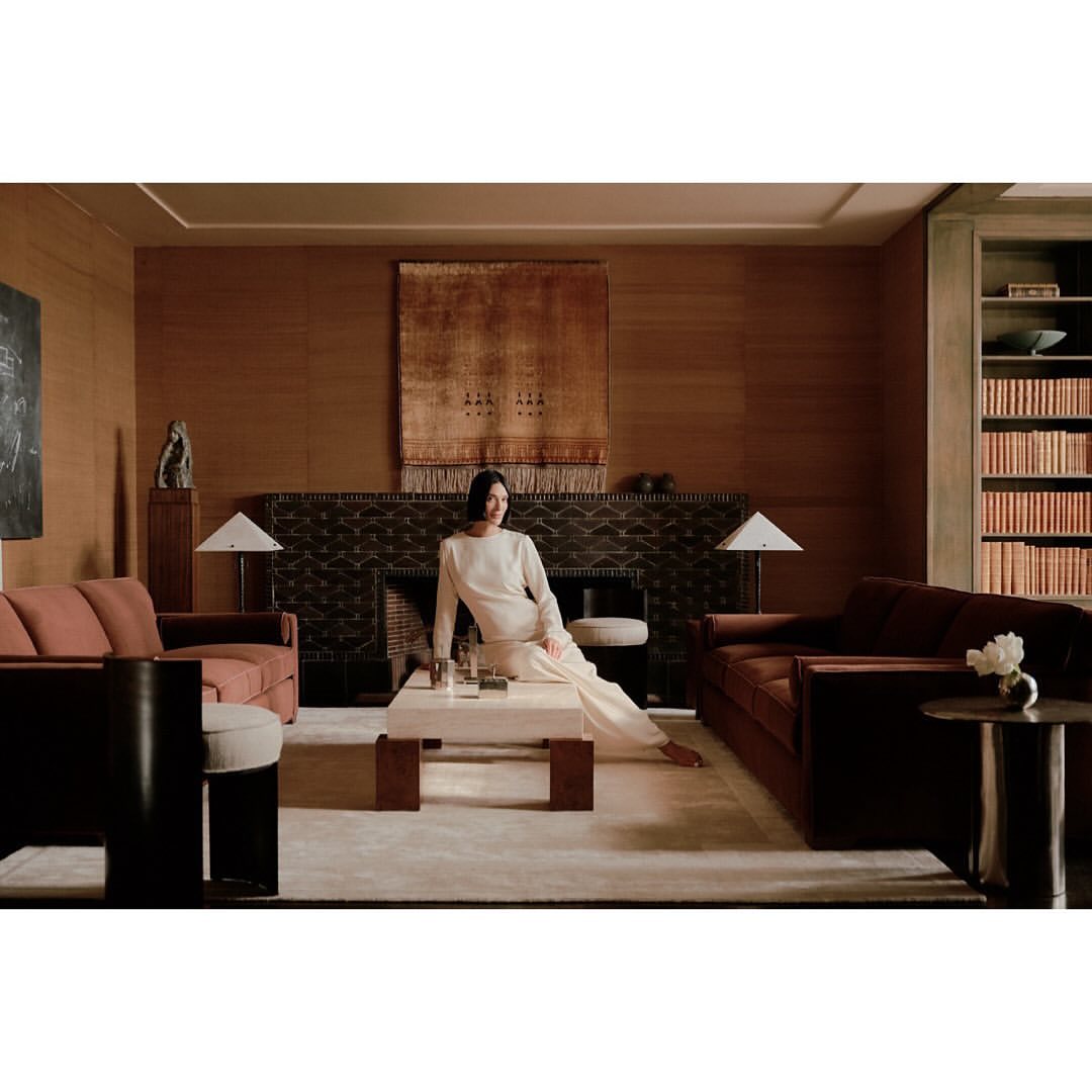 A woman in a cream-colored outfit poses in a warm, inviting living room featuring sofas, a coffee table, and an earthy color palette.