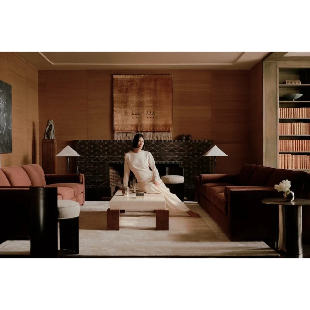 A woman in a cream-colored outfit poses in a warm, inviting living room featuring sofas, a coffee table, and an earthy color palette.