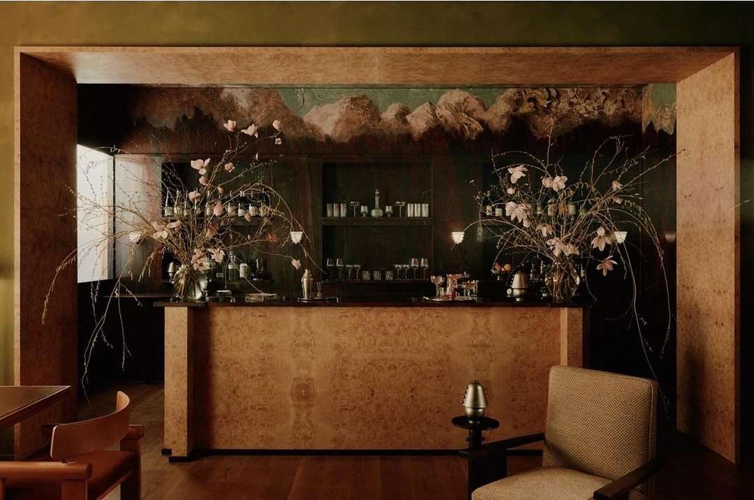 Interior view of a home bar with a flower arrangement, bottles, glasses, and shaker on a shelf, with textured walls.