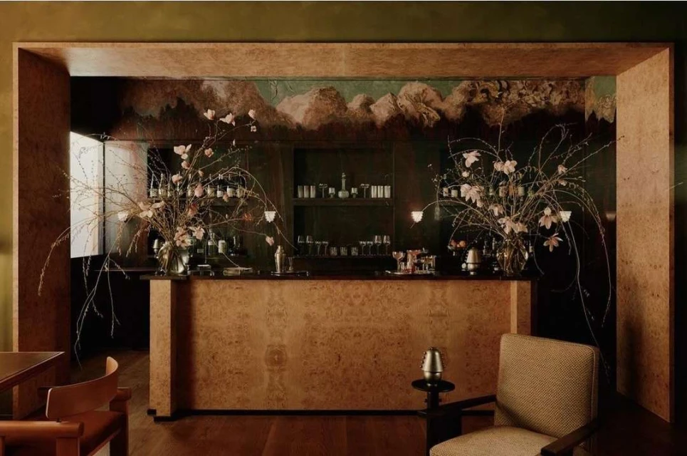 Interior view of a home bar with a flower arrangement, bottles, glasses, and shaker on a shelf, with textured walls.