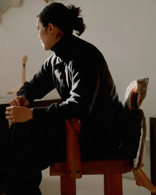 Adult male in dark turtleneck sweater sits indoors on wood chair in diffused sunlight with neutral wall backdrop, looking away.