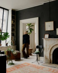 Woman in a black turtleneck and brown trousers standing in a doorway of an apartment with dark walls and a marble fireplace.