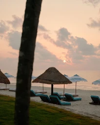A tranquil tropical beach at sunset with thatched parasols and sun loungers lining sandy shore.