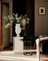 White ceramic vase with delicate light purple flowers resting on a metallic pedestal in a living room interior.
