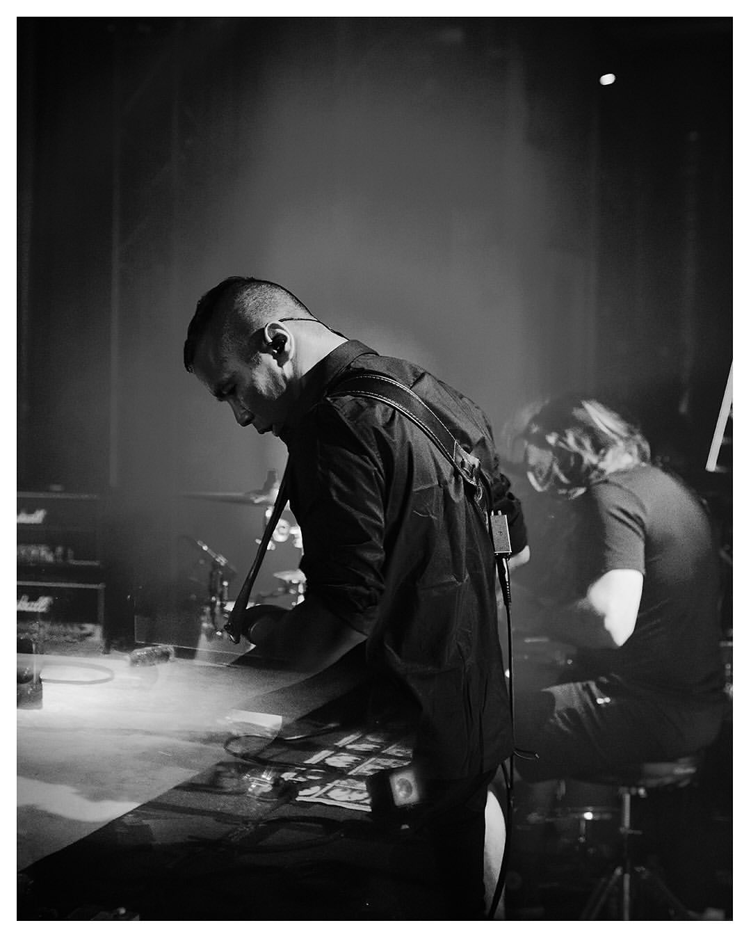 Monochrome shot of a musician in a black shirt performing on stage with a drummer in the background in a dimly lit venue.