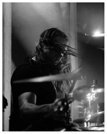 Black and white shot of a musician playing drums on stage, with long hair and a dark shirt in a dimly lit environment.