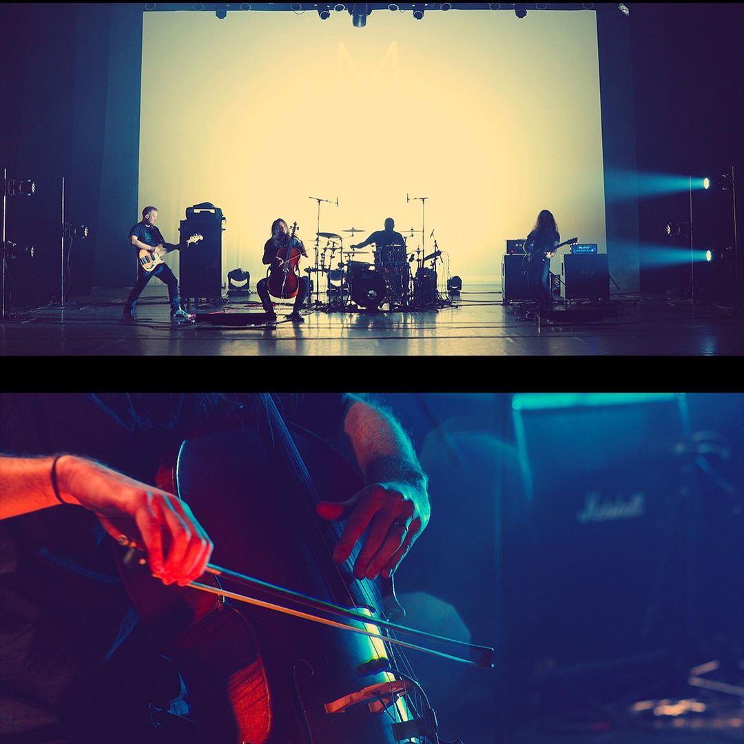 Live performance featuring a band on stage playing instruments in a brightly lit theater with a large blank screen behind them.