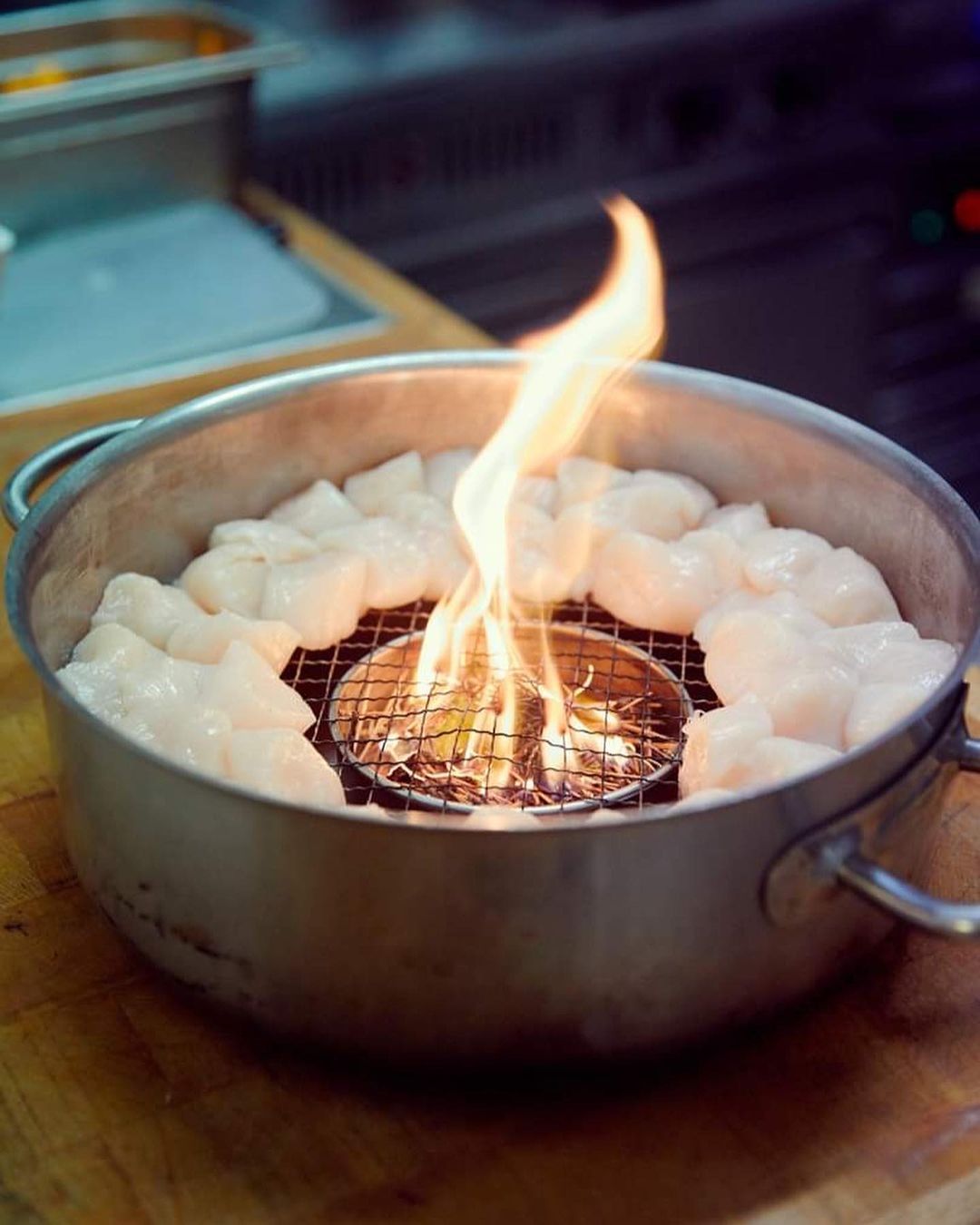 Close-up of scallops arranged around a flaming grill in a silver pot on a wooden surface, with a kitchen setting in the background.