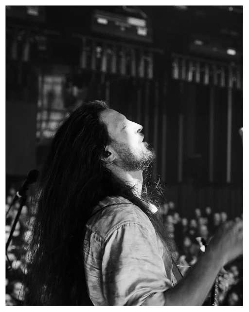 Monochrome portrait of an adult male musician with long hair, looking upwards while performing live on stage to a large audience.