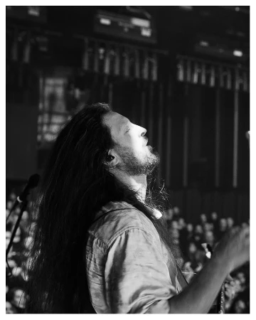 Monochrome portrait of an adult male musician with long hair, looking upwards while performing live on stage to a large audience.