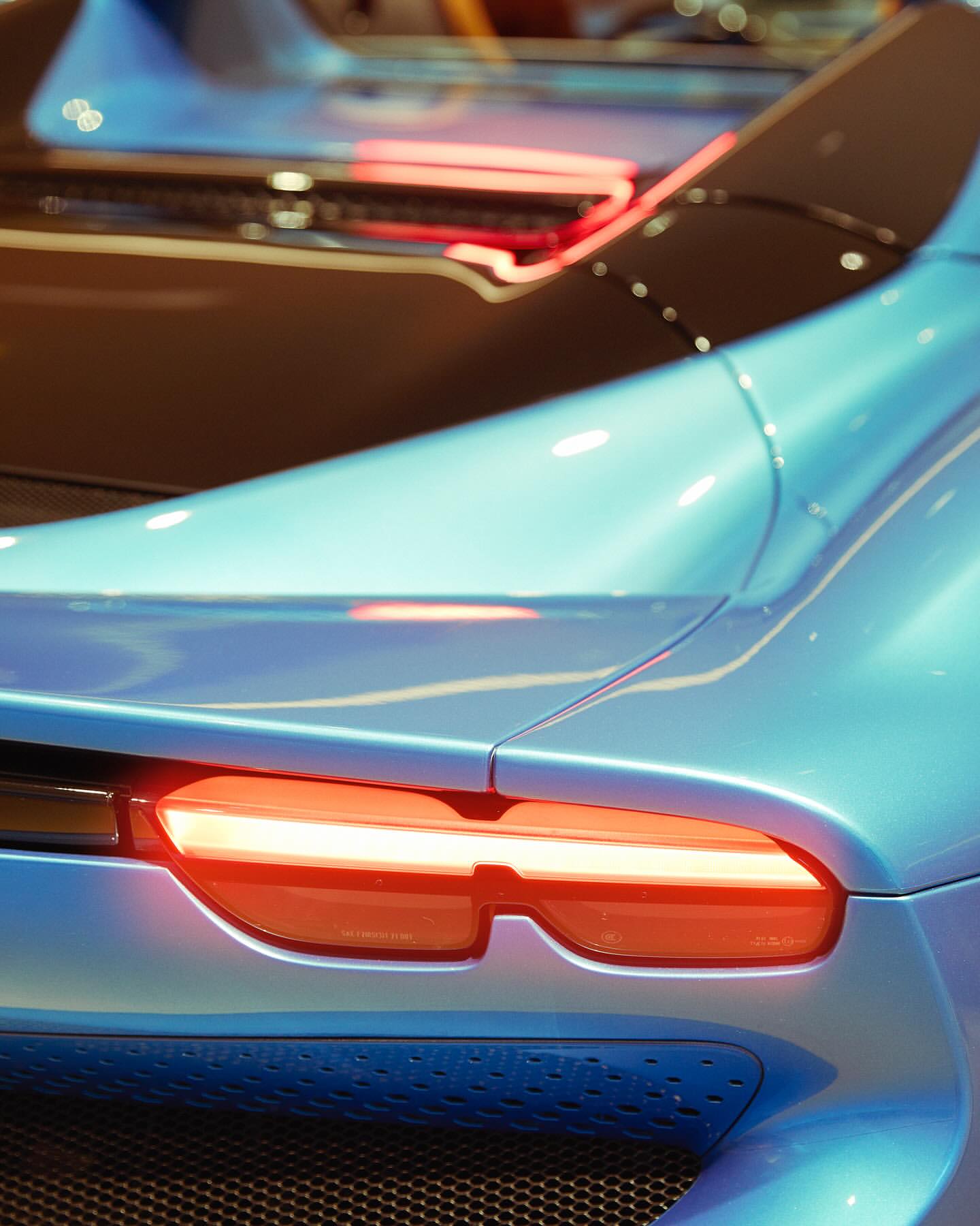 Close-up of a light-blue sports car's rear, featuring the taillights and a black spoiler. The focus is on the vehicle's design and details.