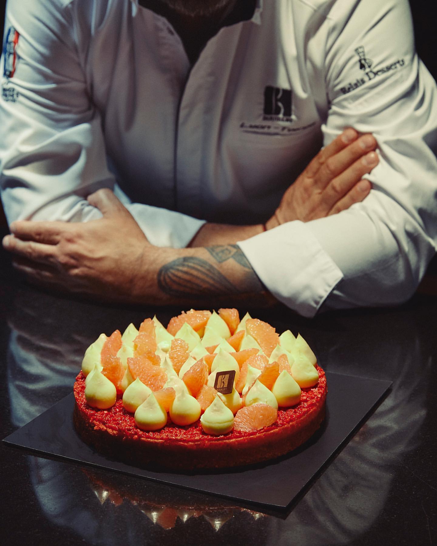 Chef poses behind a vibrant citrus tart on a black plate, showcasing its grapefruit slices, pastry cream, and glistening glaze.
