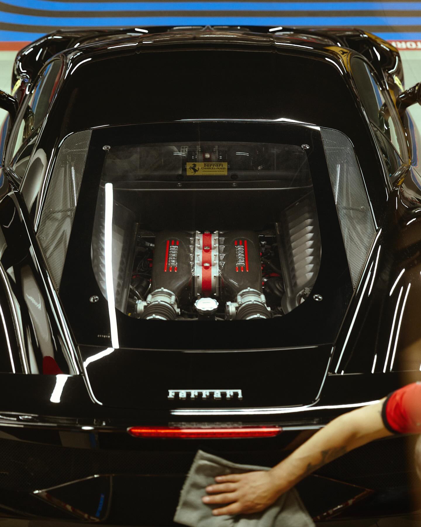 Close-up of a polished black Ferrari engine bay being cleaned, with focus on the engine and car's sleek design.
